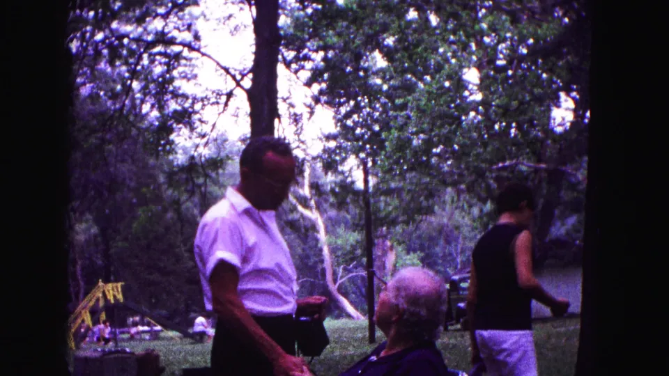 The home-movie frame shows a man with a camera, a seated older woman, and another adult moving through a wooded Galena park.