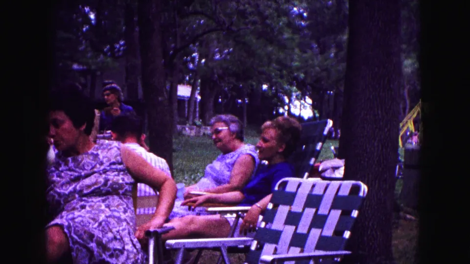 Several adults relax in folding chairs under trees, with a patterned dress and striped lawn chair visible in the 1967 park footage.