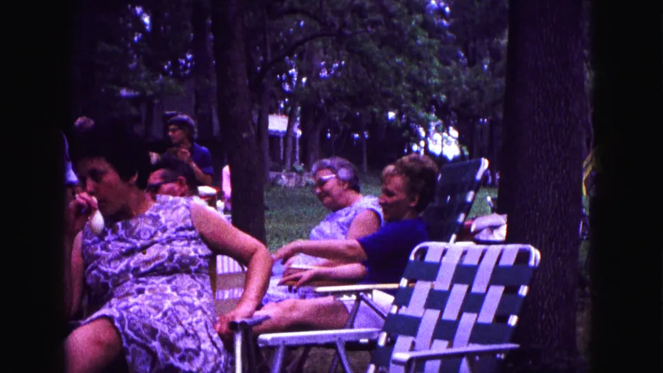 A woman in a patterned sleeveless dress sits near other women on lawn chairs in 1967 Galena home-movie footage.