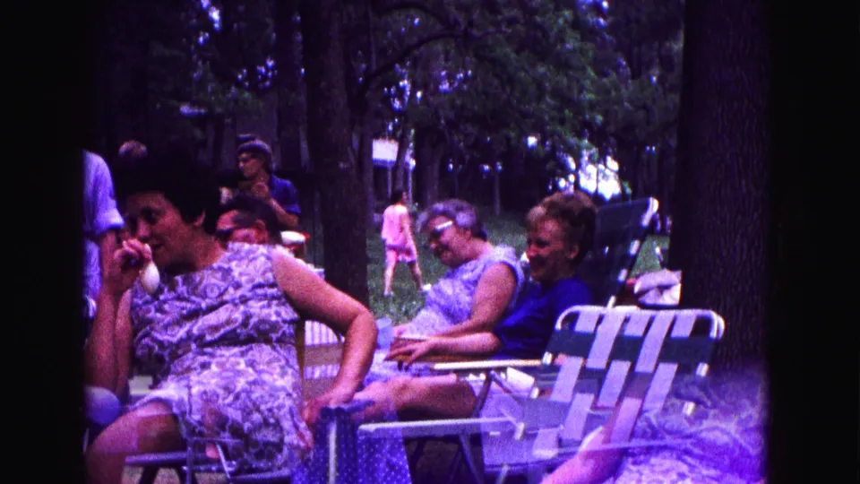 Women remain seated in lawn chairs under trees, with a distant figure visible in the background, in 1967 Galena footage.