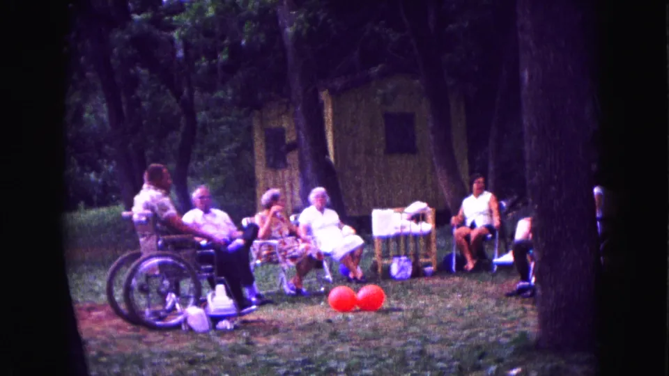 1967 backyard party frame with a wheelchair user seated among relatives in lawn chairs near red balloons.