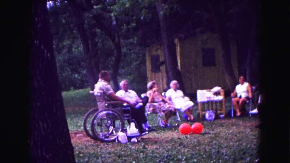 Galena, Illinois home-movie frame of adults seated in lawn chairs under trees during a family birthday gathering.