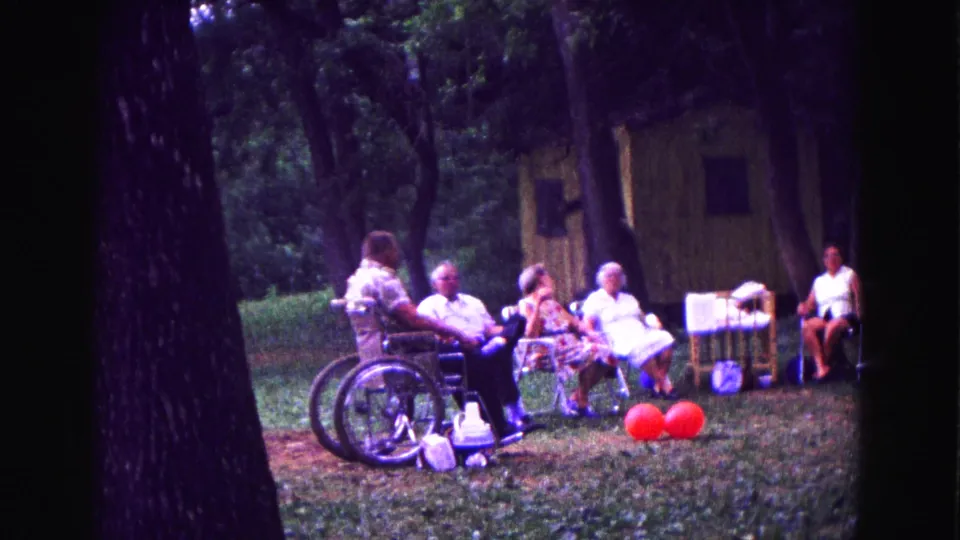 1967 Galena, Illinois home-movie frame of older adults seated outdoors in a wooded area with a wheelchair, chairs, red balloons, and a small rustic structure.