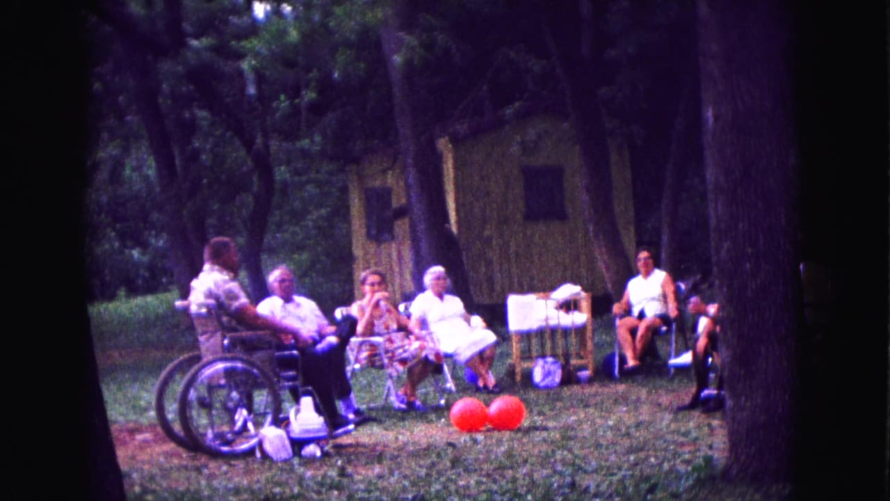 Older adults in 1967 Galena, Illinois sit and talk outdoors beside a wheelchair, red balloons, folding chairs, trees, and picnic supplies.