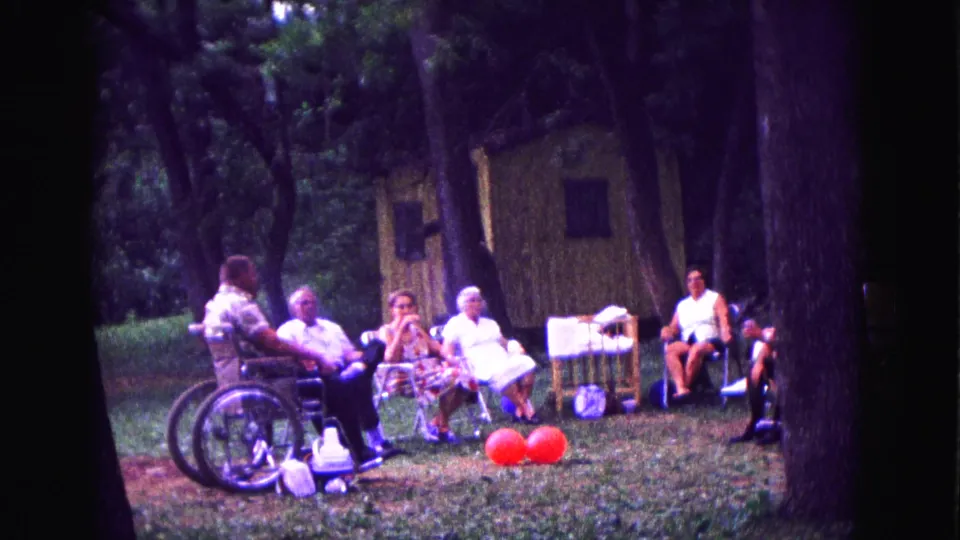 Older adults in 1967 Galena, Illinois sit and talk outdoors beside a wheelchair, red balloons, folding chairs, trees, and picnic supplies.