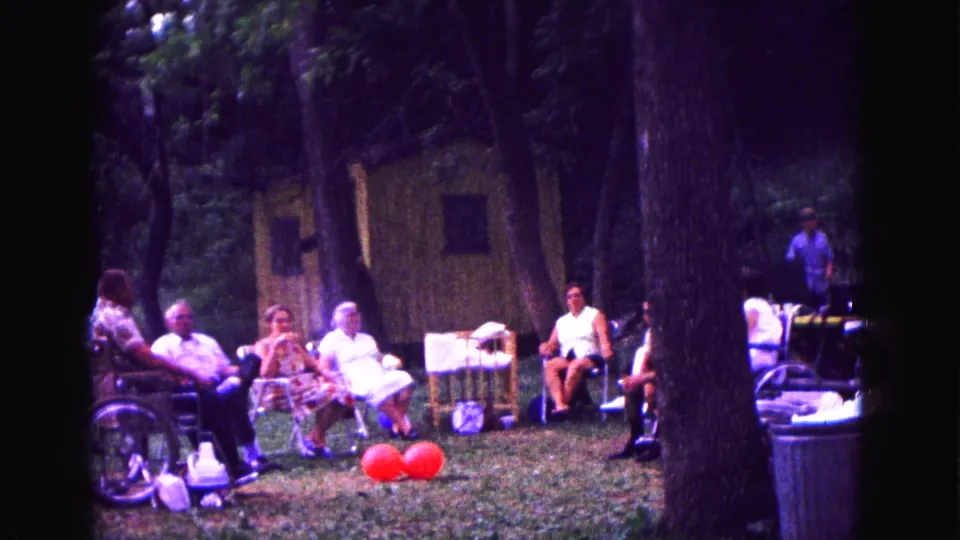 1967 Galena, Illinois frame showing older adults seated outdoors near a wheelchair, red balloons, picnic gear, trees, and a partly visible standing figure.