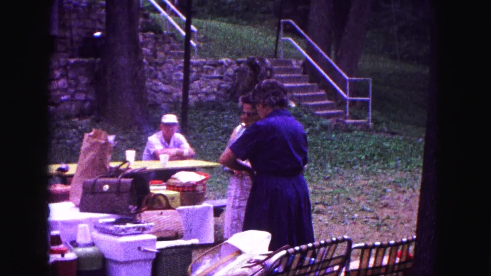 1967 Galena, Illinois home-movie frame of picnic supplies, a standing woman, and a seated man in a hat near stone steps in a wooded setting.