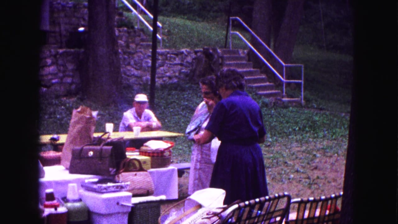 1967 Galena, Illinois 8mm frame showing women near picnic tables while a man in a white cap sits beyond the food and bags.
