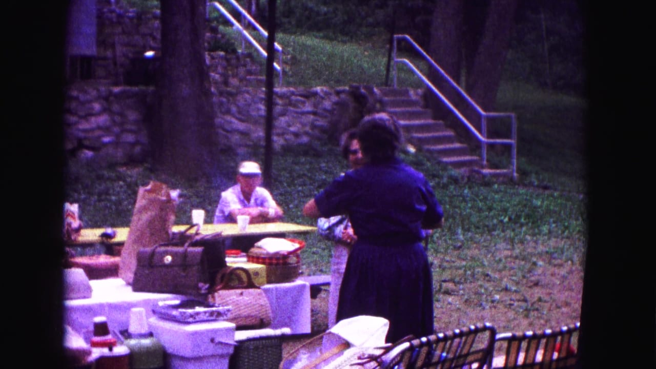 Outdoor 1967 Galena picnic scene with adults talking beside tables loaded with bags, baskets, coolers, and cups under trees.