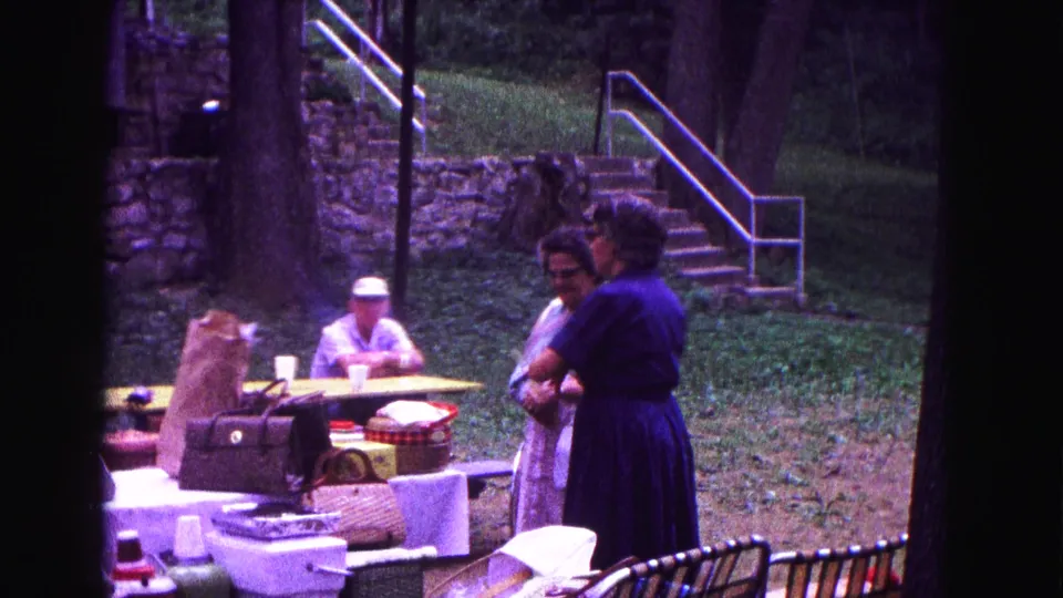 1960s home-movie frame from Galena showing adults gathered around picnic tables with food containers, bags, trees, and stone steps.