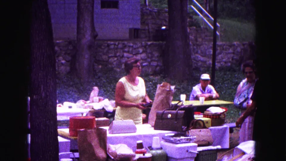 1967 Galena picnic frame with a woman arranging items at an outdoor table, another woman at right, and a seated man behind.