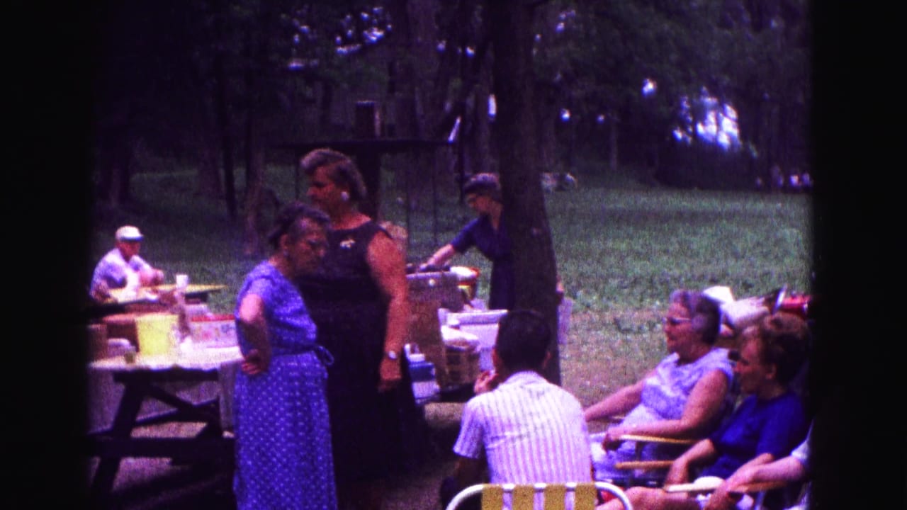 Adults at a 1967 Galena, Illinois picnic gather near covered tables, folding chairs, and trees in 8mm home-movie footage.