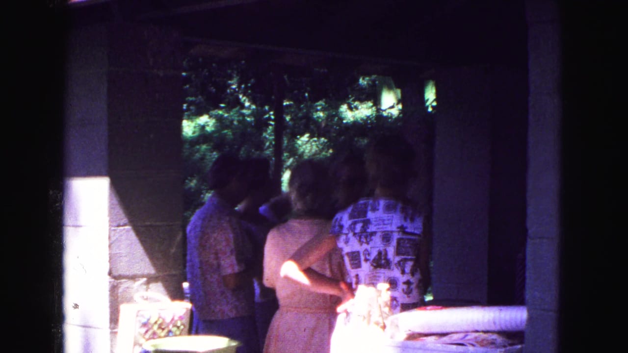 Adults stand in porch shade near a table, with bright sun on concrete blocks and trees beyond.