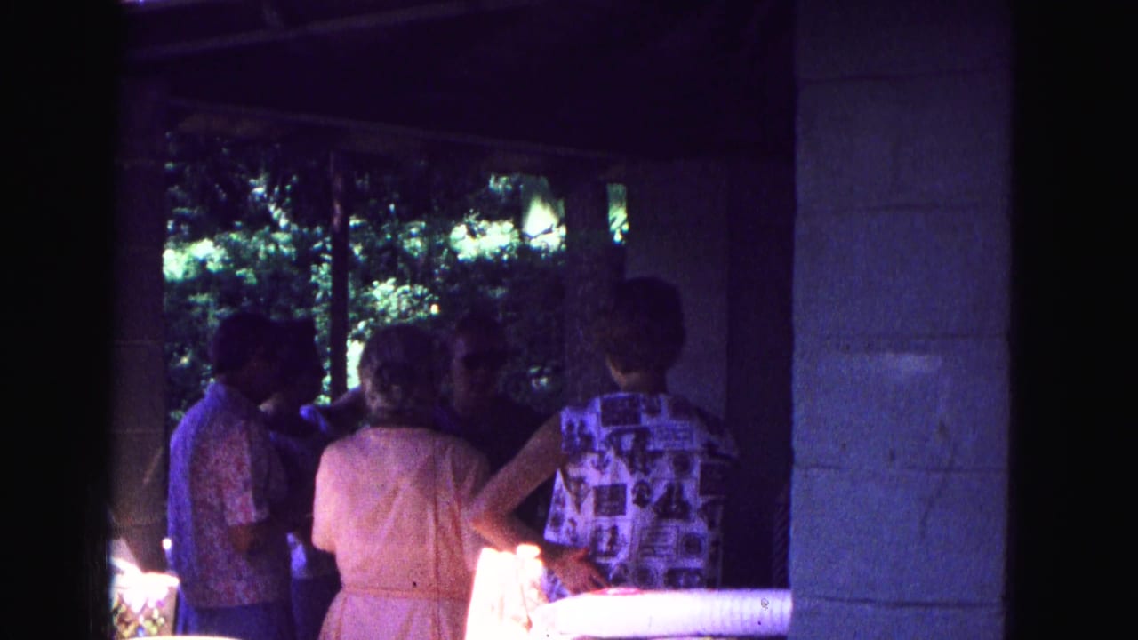 Group of adults stands close together under a porch, with a woman in a pale dress facing away.