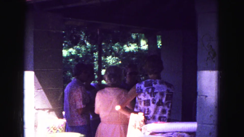 The 8mm frame shows adults clustered under a shaded porch at a 1967 Galena picnic, with bright sun striking the concrete wall.