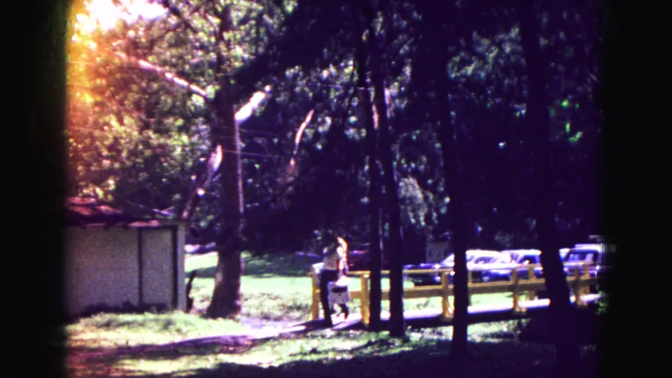 In the 1967 Galena park scene, a man walks through shade near the yellow fence, with parked cars beyond the trees.