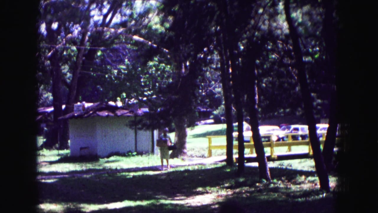 Woman walking near a small white shed in a wooded park with a yellow fence behind her.
