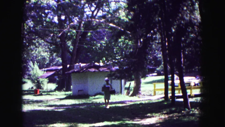 1967 Galena, Illinois 8mm frame of a woman walking toward the camera with a lunch pail in a wooded park setting.