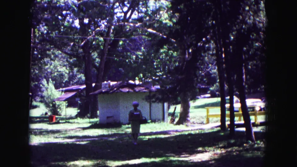 1967 Galena, Illinois home-movie frame showing the woman centered in the park, carrying a lunch pail as trees and a small shed fill the background.