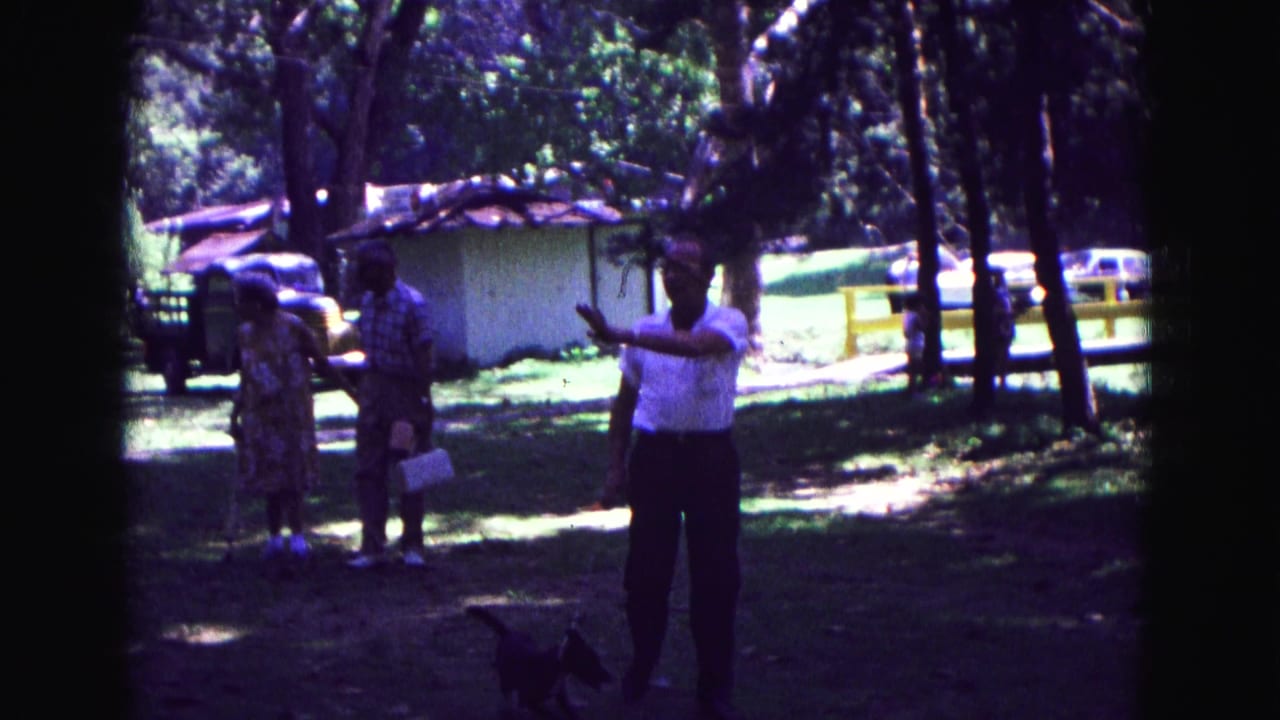 1967 Galena, Illinois 8mm frame showing a man in a white shirt with a small leashed dog as two adults stand in a shaded grassy area.