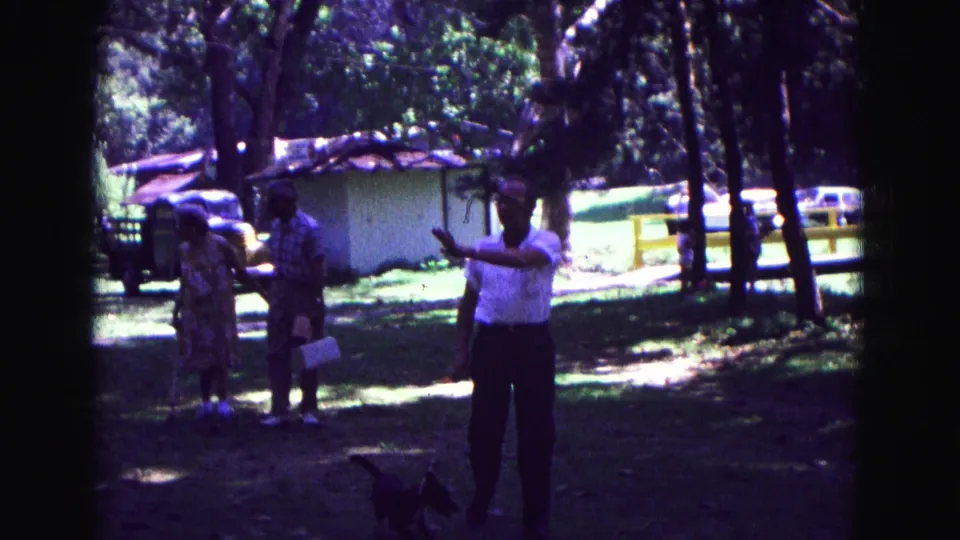 1967 Galena, Illinois 8mm frame showing a man in a white shirt with a small leashed dog as two adults stand in a shaded grassy area.
