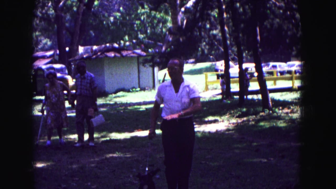 1967 Galena, Illinois home-movie frame with a man in a white short-sleeved shirt walking toward the camera in a grassy, tree-shaded area.