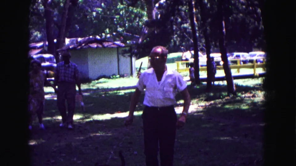 1967 Galena, Illinois frame showing a man in a white shirt walking across grass, with two adults, trees, cars, and a shed behind him.