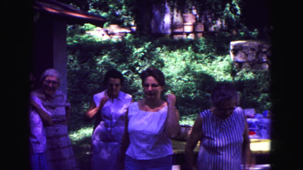 Five adult women stand on a shaded porch with bright greenery behind them.