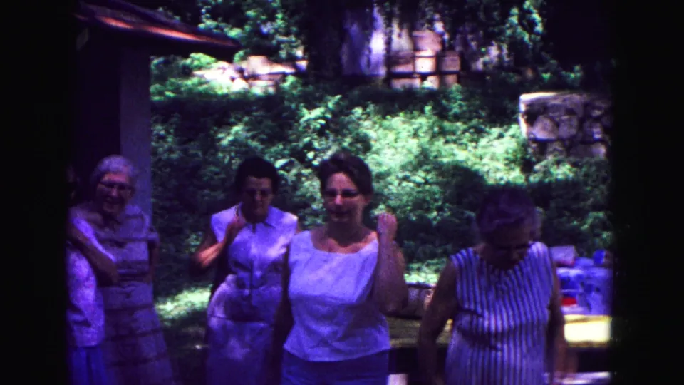 Five adult women in summer clothing stand and chat on a shaded porch in Galena, Illinois, in 1967 home-movie footage.