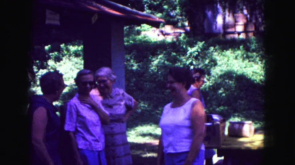 Adult women continue a porch conversation in 1967 Galena home-movie footage, with bright garden greenery in the background.