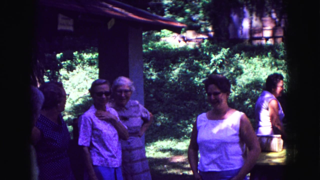 Women stand and talk beneath a shaded porch with trees behind them.