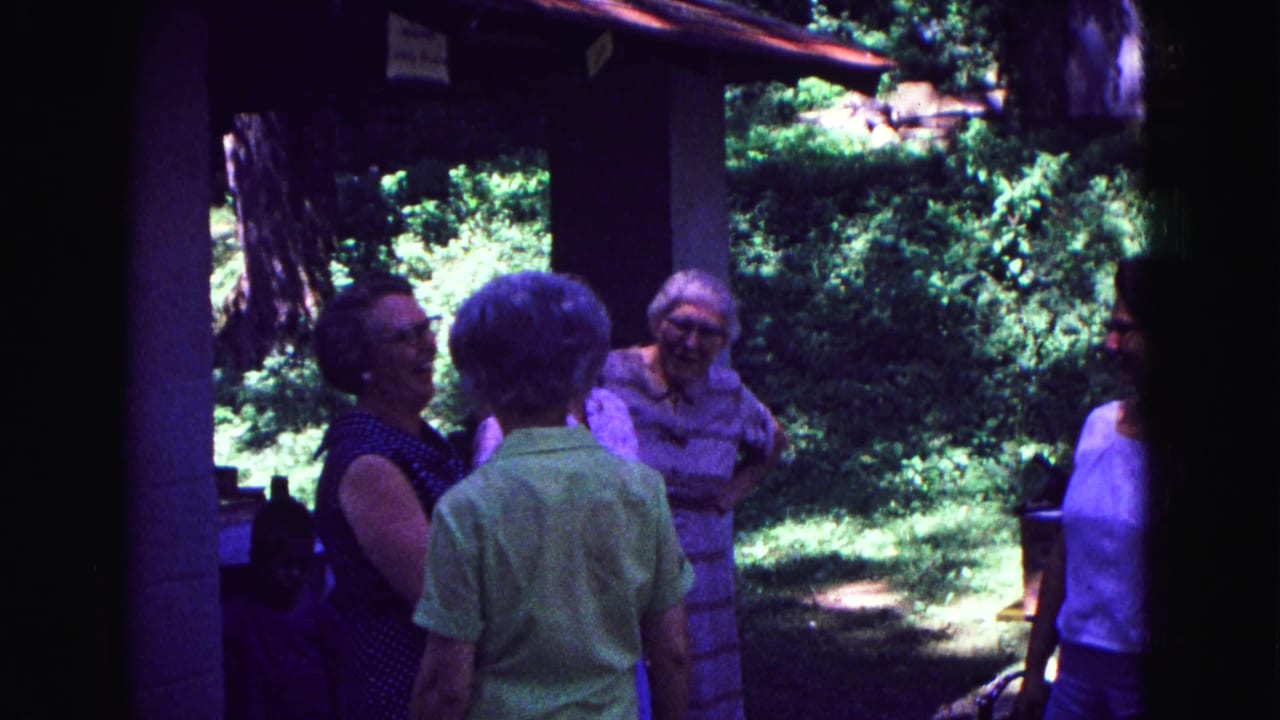 Several women in summer clothing converse near porch posts and trees in the 1967 Galena home-movie scene.