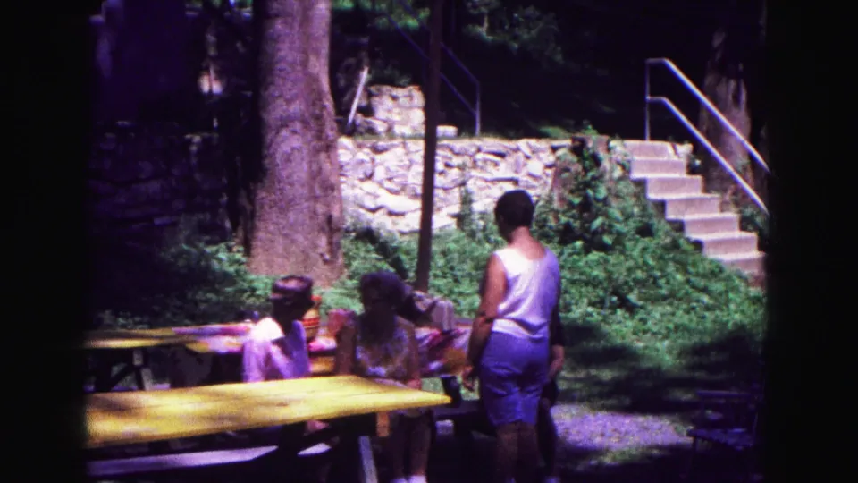 Women sit and stand around yellow picnic tables in Galena, with a large tree, stone wall, and stairs behind them.