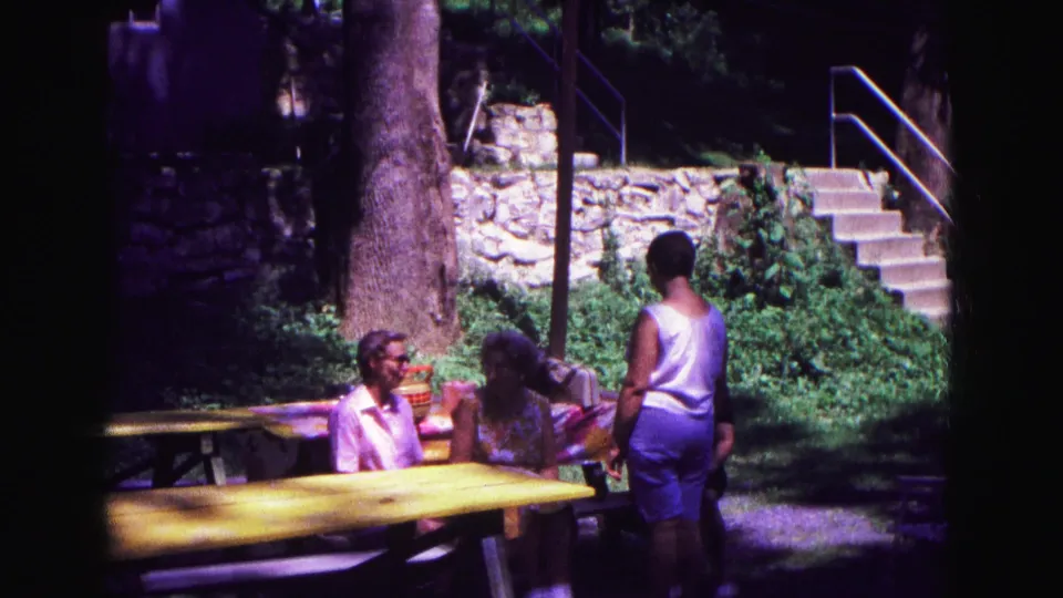 A standing woman faces seated women at yellow picnic tables during a 1967 Galena outdoor stop.