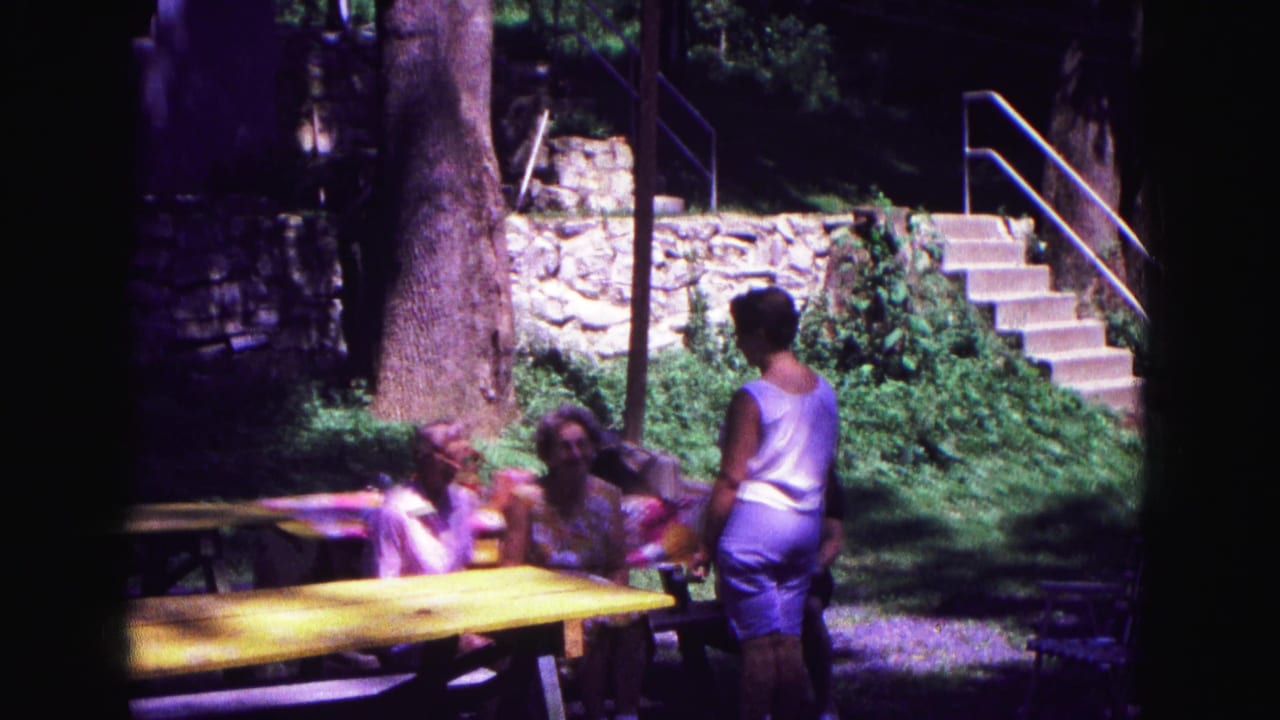 1967 Galena outdoor frame with adults conversing beside a yellow picnic table under trees near a stone wall and stairs.