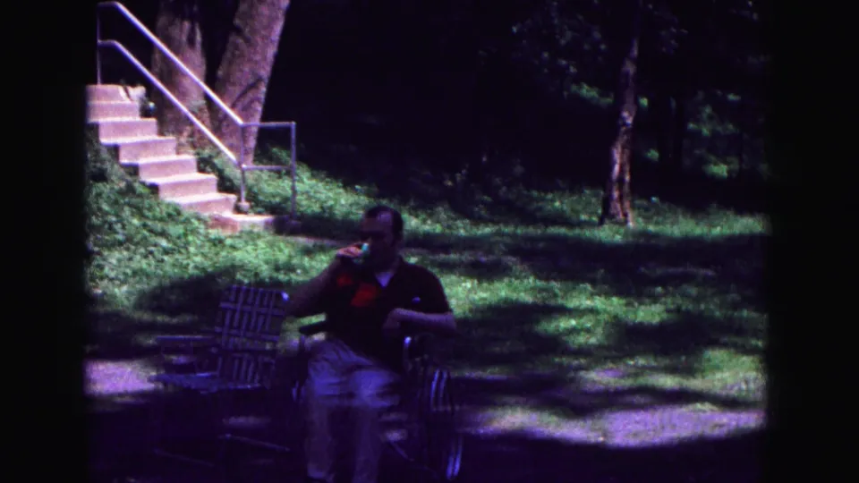 1967 Galena home-movie frame showing a seated man drinking in a lawn chair with stairs, grass, and trees behind him.