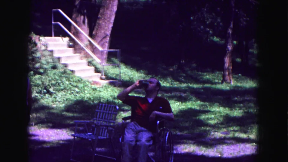 1967 Galena frame of a seated man drinking outdoors, with lawn chairs, stairs, trees, and dappled sunlight visible.