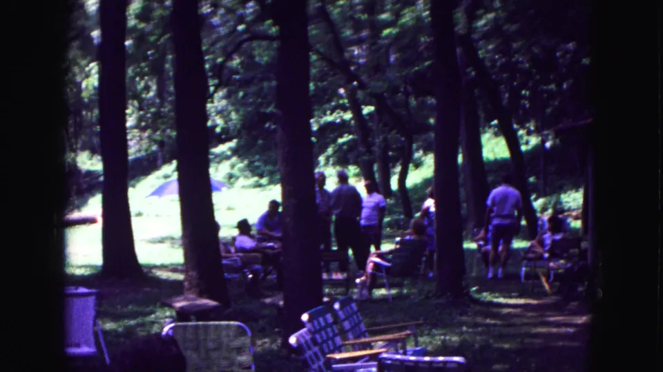 Adults sit and stand among trees in a shaded Galena picnic area, with folding chairs and a grassy clearing visible.