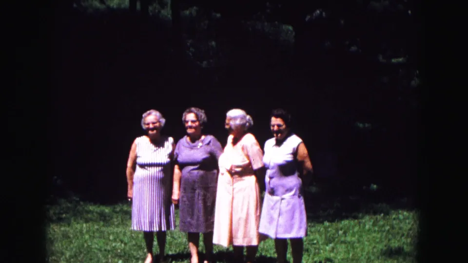 The 1967 home-movie frame shows four older women smiling shoulder to shoulder on grass, with a dark wooded backdrop.