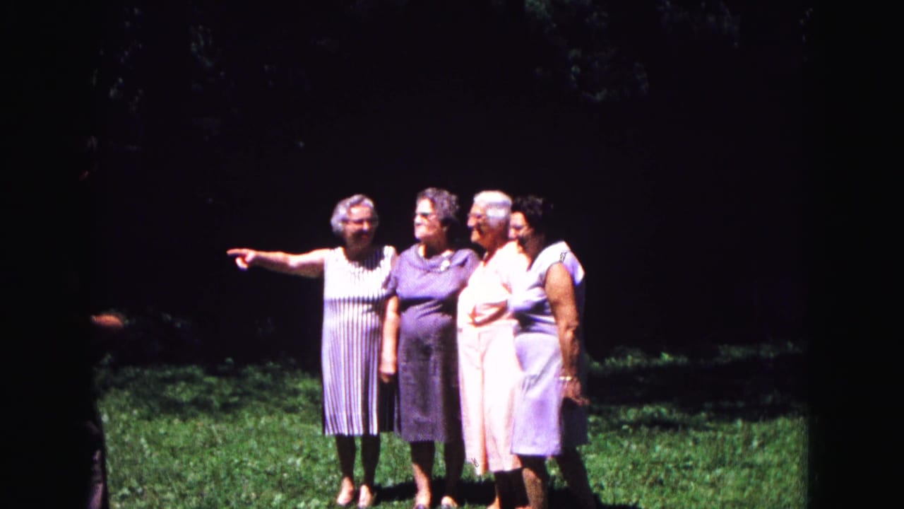Four older women in summer dresses stand on a lawn, with one woman pointing off-camera.