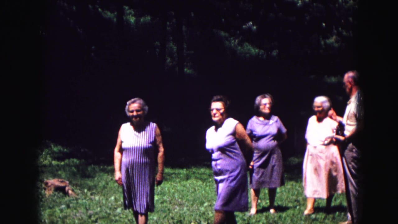 Older women walk across a shaded lawn while a man stands at the right edge.