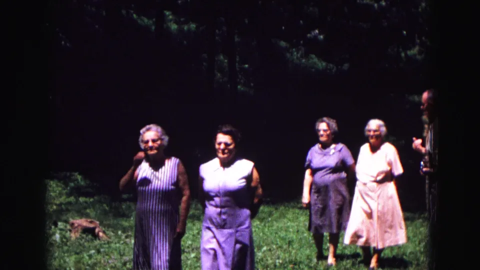 Two older women in sleeveless dresses approach the camera in a Galena yard, with two more women and a man partly visible behind them.