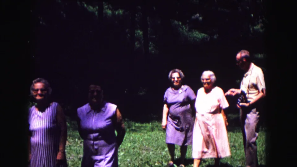 1967 Galena home-movie frame showing several adults standing on grass in dappled shade, with trees behind them.