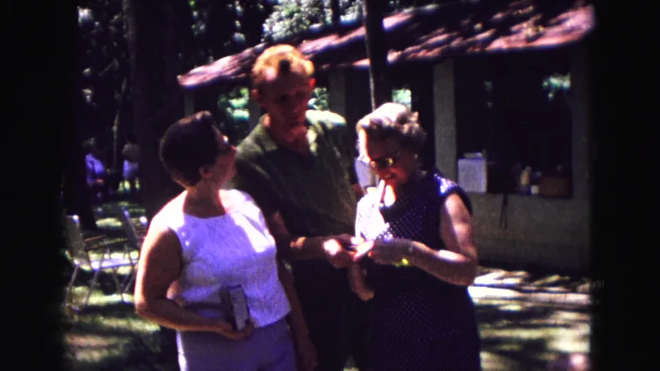 1967 Galena frame showing a younger man offering a small snack to an older woman beside a rustic building.