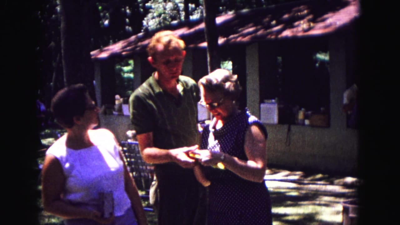Three adults stand outdoors as a woman in a polka-dot dress opens a small gift.