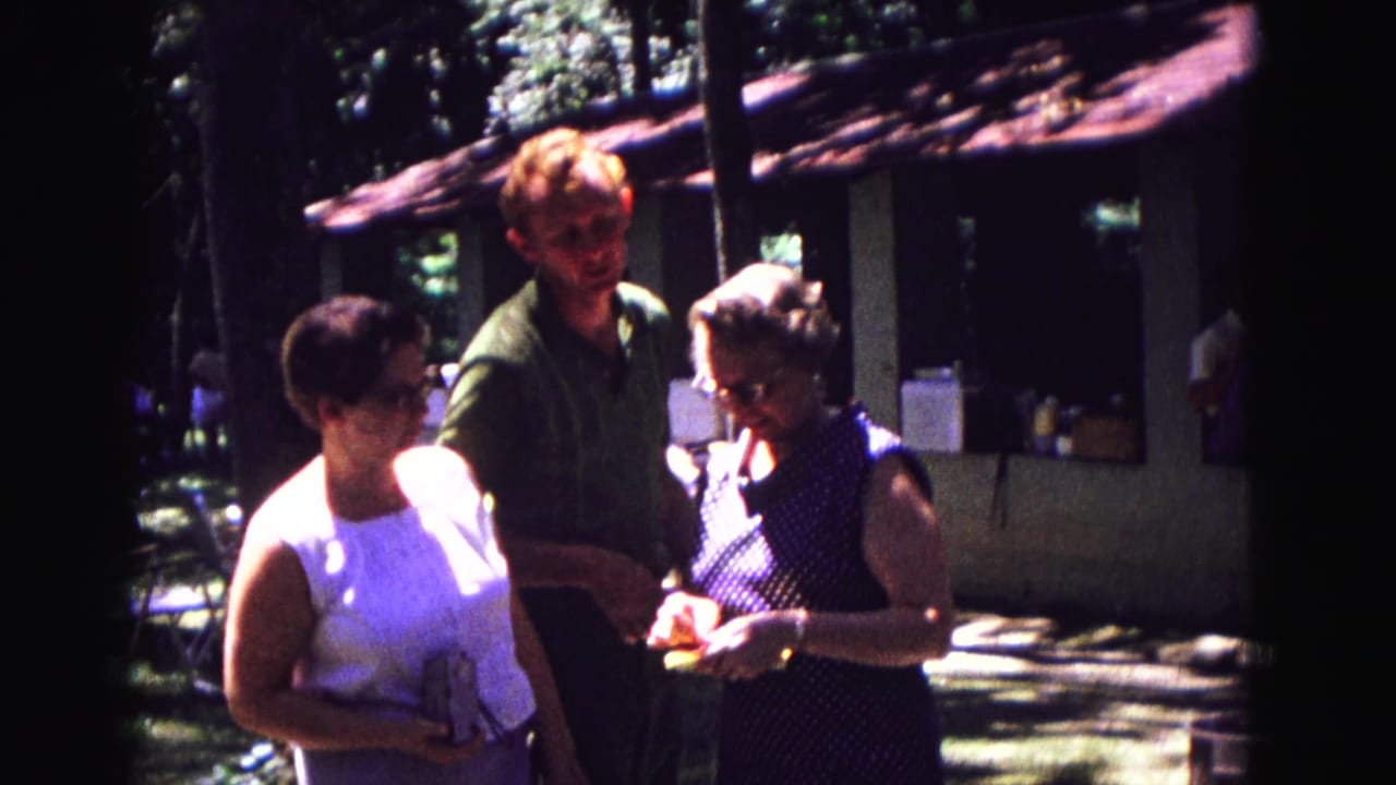 Adults at a 1967 Galena outdoor family gathering stand together while a woman in a polka-dot dress handles a small gift.