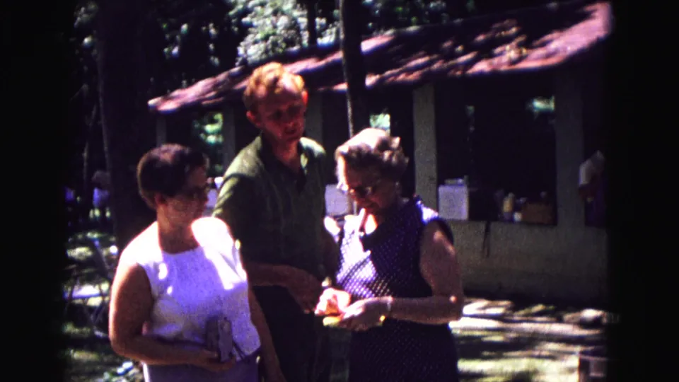 Adults at a 1967 Galena outdoor family gathering stand together while a woman in a polka-dot dress handles a small gift.