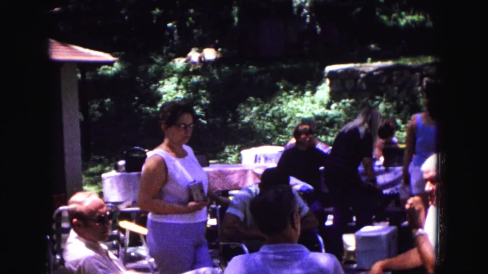 Backyard party guests in Galena, Illinois, with a woman standing near tables and adults seated under bright 8mm sunlight.