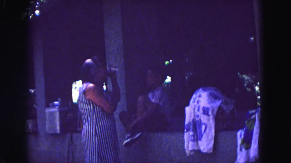 1967 Galena, Illinois 8mm frame showing a woman in a striped dress holding a drink near a pillar as others sit in shadow.
