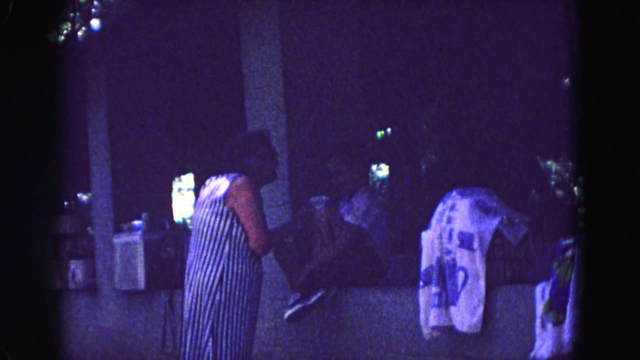 1967 Galena, Illinois home-movie frame with a standing woman, seated adults, pillars, and draped fabric in low purple-tinted light.